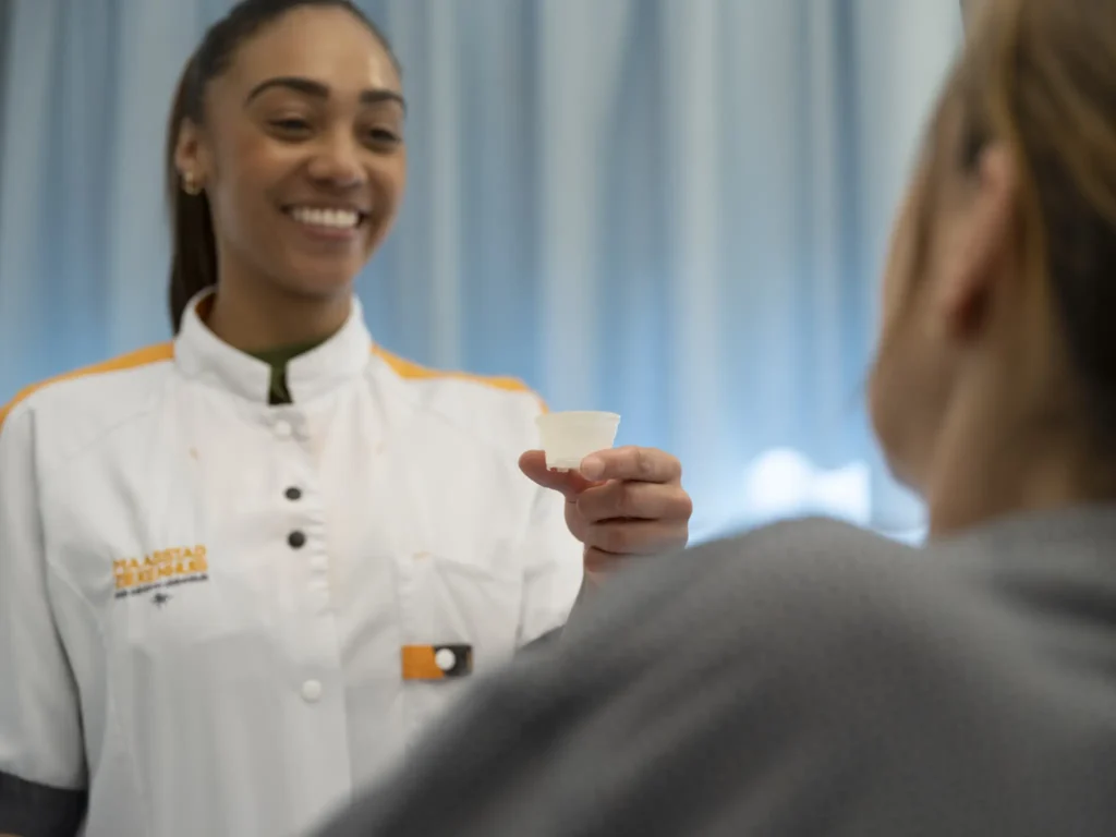 Nagespeelde foto. Een verpleegkundige op de afdeling houdt lachend een medicijnbekertje van cellulose omhoog